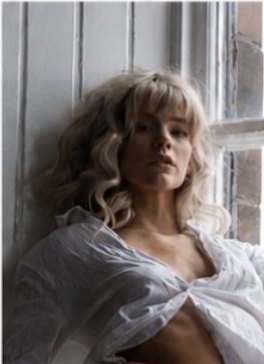 Charlotte S seated by a window in a sheer white shirt